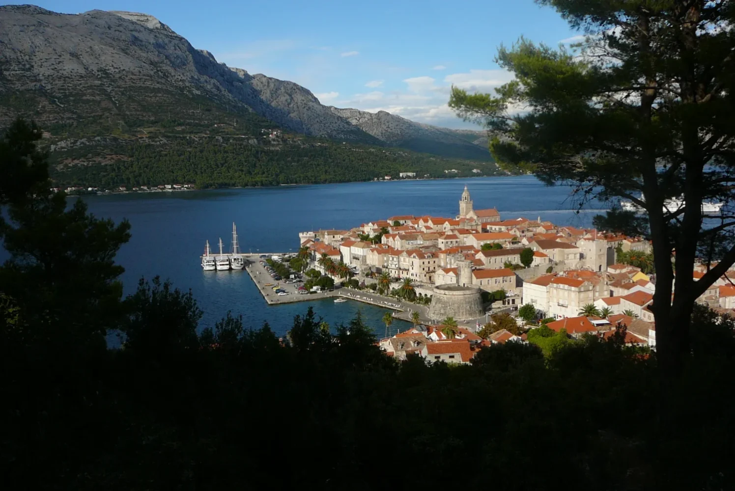 Panoramic view of historic Korčula Old Town with red tiled roofs, stone city walls, and ships anchored in the Adriatic harbor.