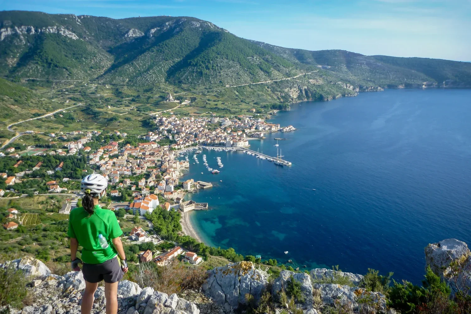 Female cyclist in green helmet standing on a rocky viewpoint looking down at a coastal village bay and deep blue Adriatic sea on Vis Island.