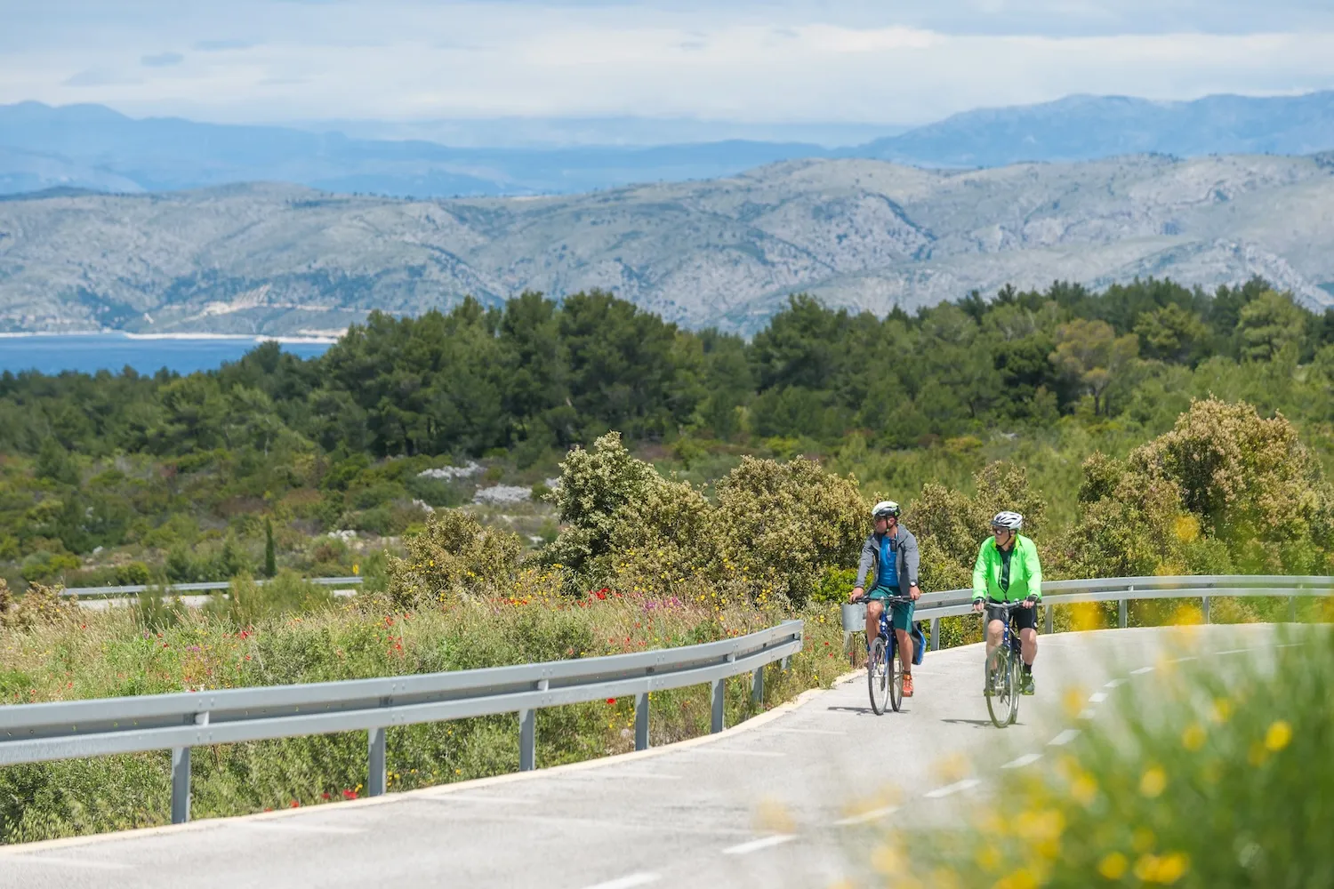 Two cyclists with helmets and panniers riding e-bikes on a scenic paved coastal road overlooking the Dalmatian mountains and Adriatic Sea.