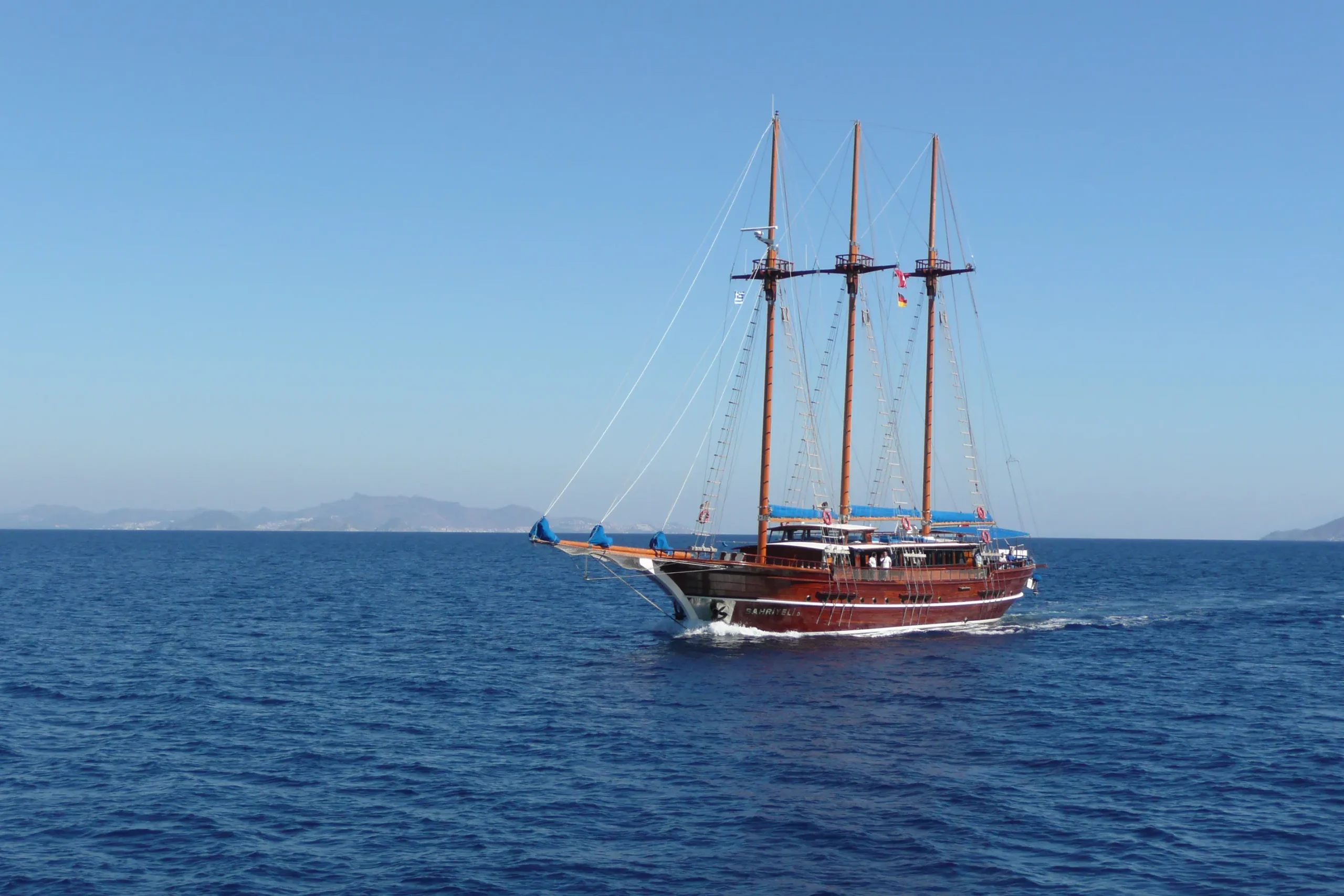 The traditional three-masted wooden motor yacht Erato cruising gracefully across deep blue waters along the Greek island coastline.
