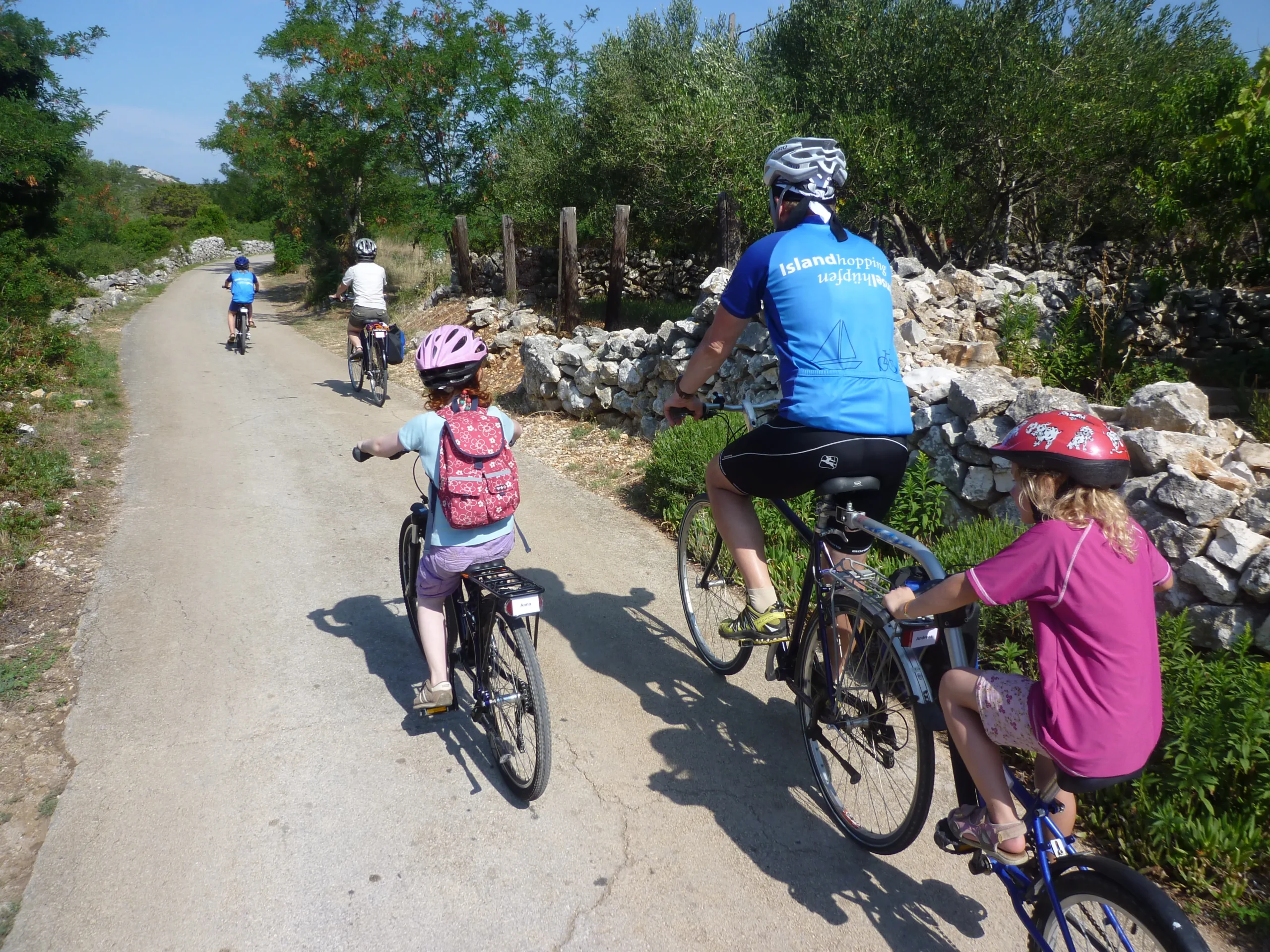 Family riding bicycles and a trailer bike along a quiet paved countryside path during a guided multi-adventure tour in Greece.