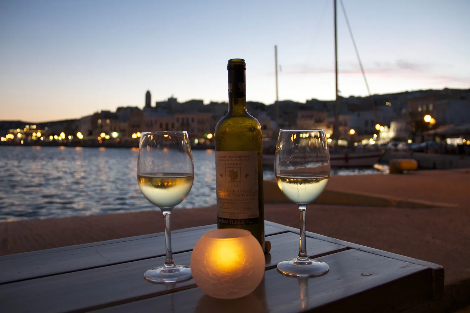 Two glasses of white Greek wine and a candle lit on a seaside table overlooking a illuminated harbor town at dusk.