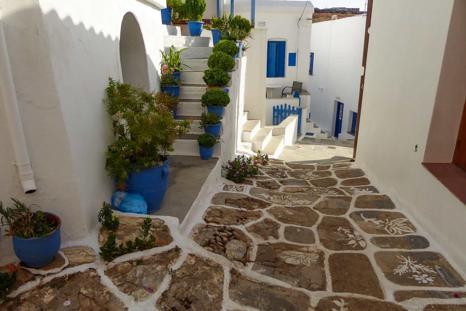 Picturesque narrow street in Chora, Kythnos Island, featuring stone-paved paths with painted floral motifs and blue potted plants.