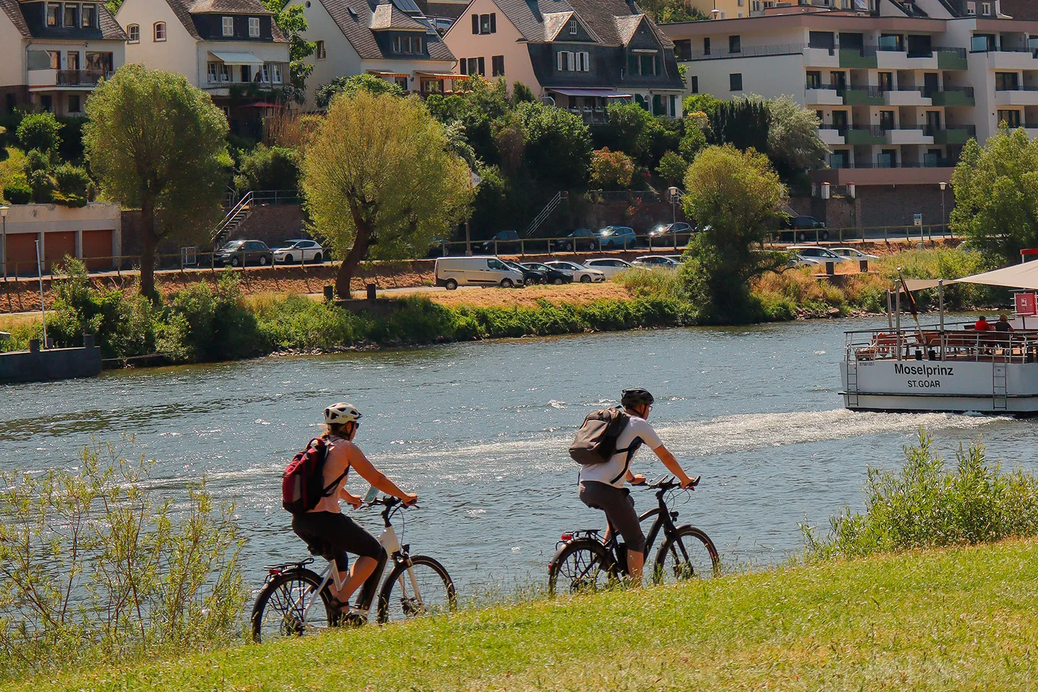 Two cyclists riding along a scenic riverside path with houses and greenery